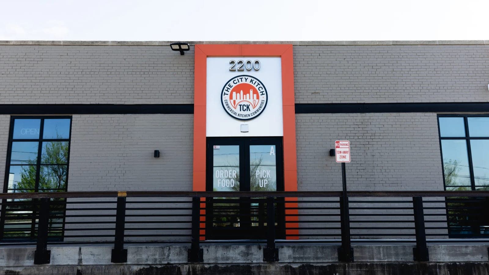 The City Kitch at 2200 Thrift Road — a grey brick building with an orange entrance bay, the TCK roundel above the door, and Order Food and Pick Up marked on the glass