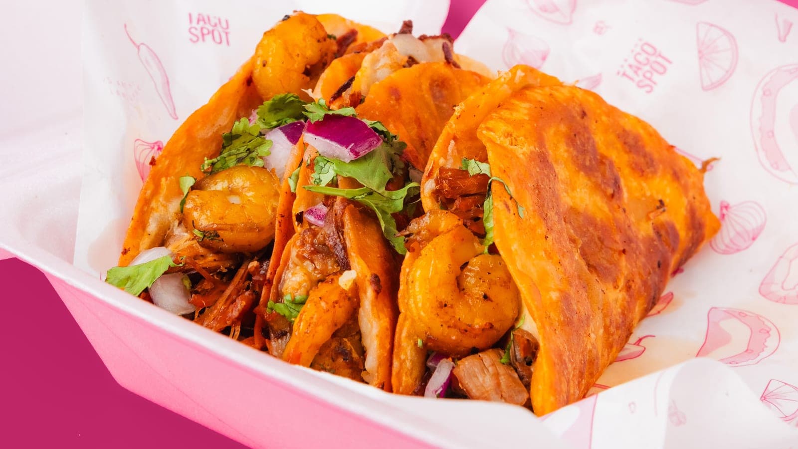 Four tacos — shrimp, al pastor and birria — in a Mariah's Taco Spot takeout container, with a hand squeezing lime over them