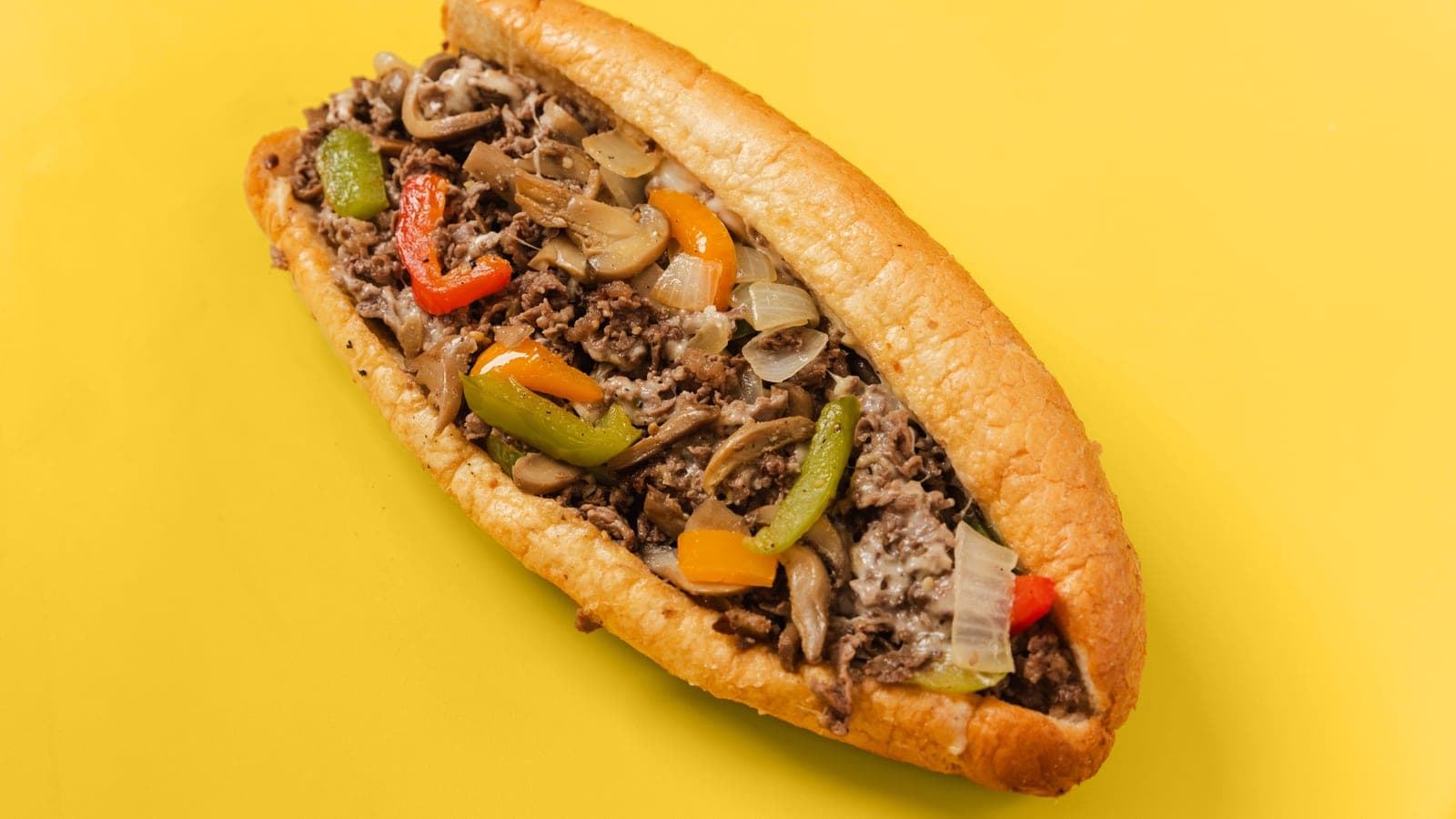 A Queen City Cheesesteaks ribeye cheesesteak with peppers, mushrooms and onions on a bright yellow background