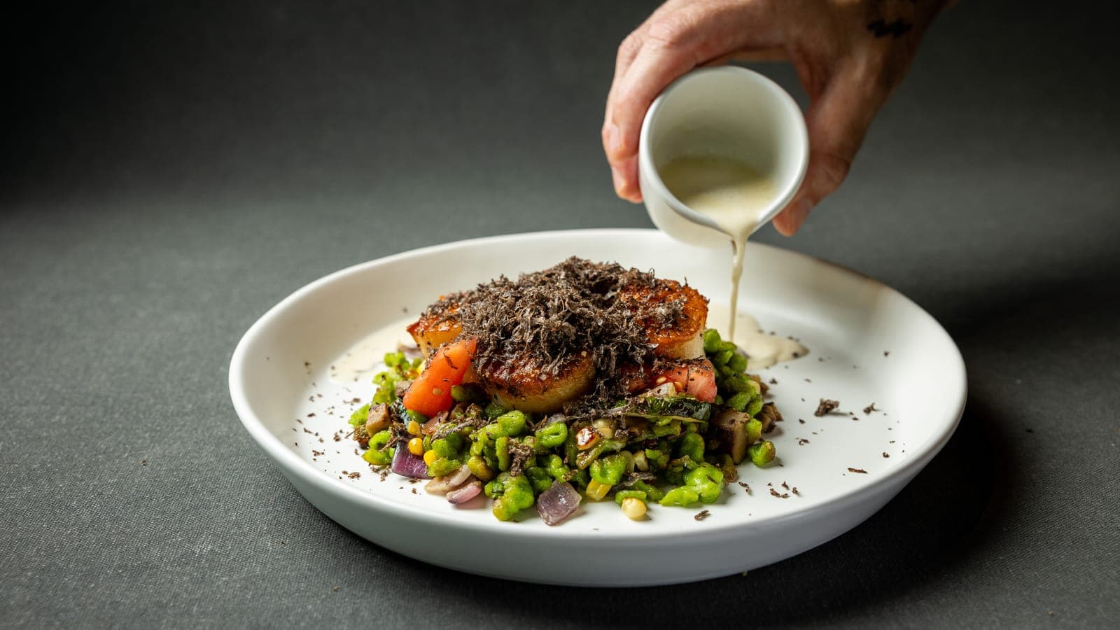 A seared scallop under a mound of shaved black truffle, on green pea spaetzle with corn and tomato, as a jug of sauce is poured over it