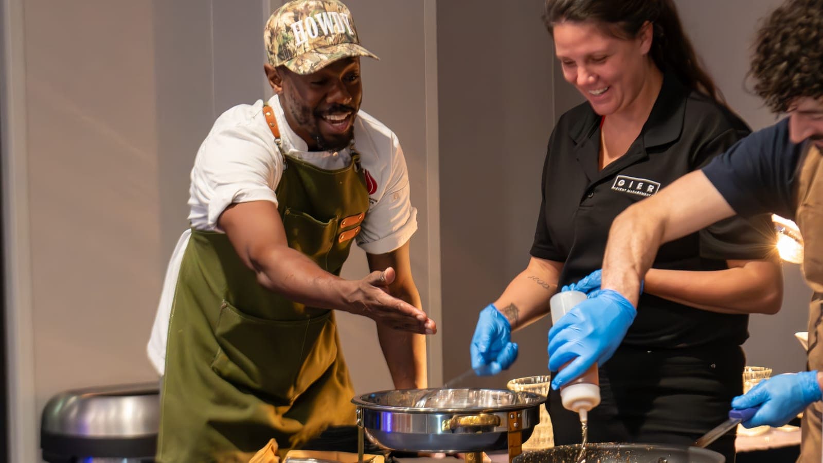 Chef William Williams laughing in a green apron as he works a chafing dish on a catering service line beside event staff