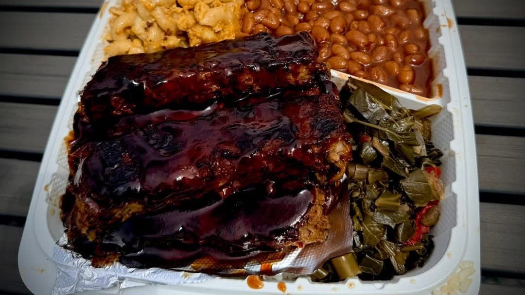 A plant-based Ribbz plate in a takeout clamshell, glazed in barbecue sauce with macaroni cheese, baked beans and collard greens
